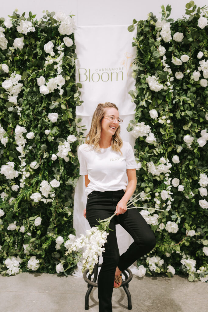 Owner is smiling and holding flowers in front of the floral wall